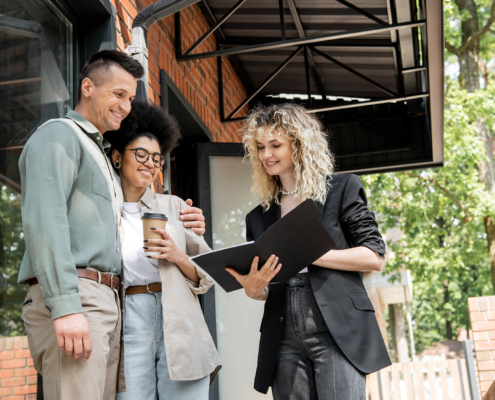 Smiling Real Estate Agent Showing Documents To Interracial Couple With Paper Cup Near Cottage